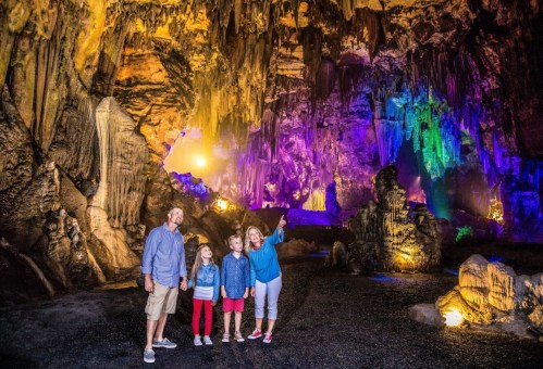A family stands in a colorful cave, illuminated by vibrant lights, surrounded by stunning rock formations.