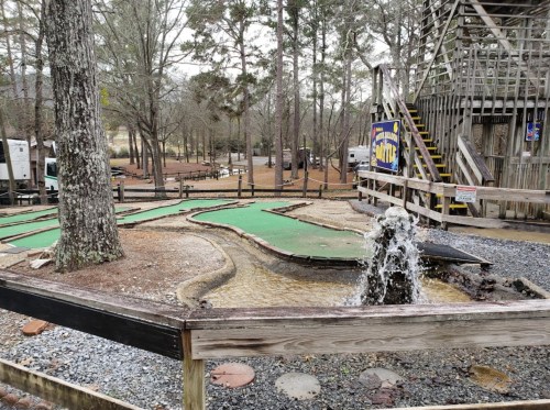 A mini-golf course with green turf, a small fountain, and trees in a wooded area, featuring wooden structures nearby.