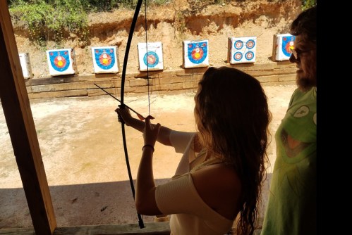 A person aims an arrow at targets while another person observes from the side at an archery range.