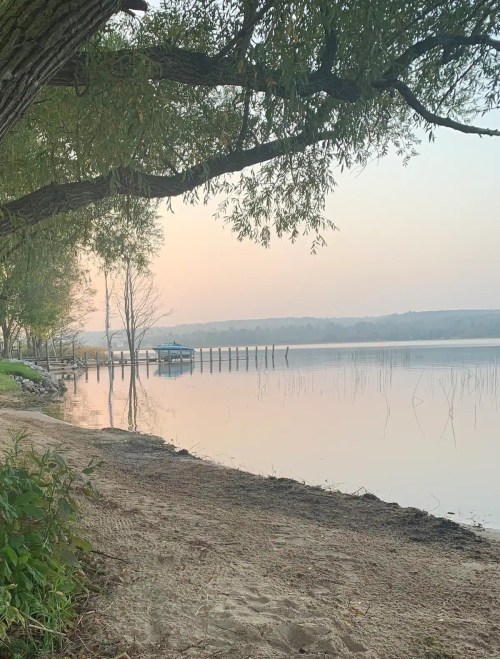 Serene lakeside view at dawn, with a sandy shore, trees, and a small boat docked in the calm water.