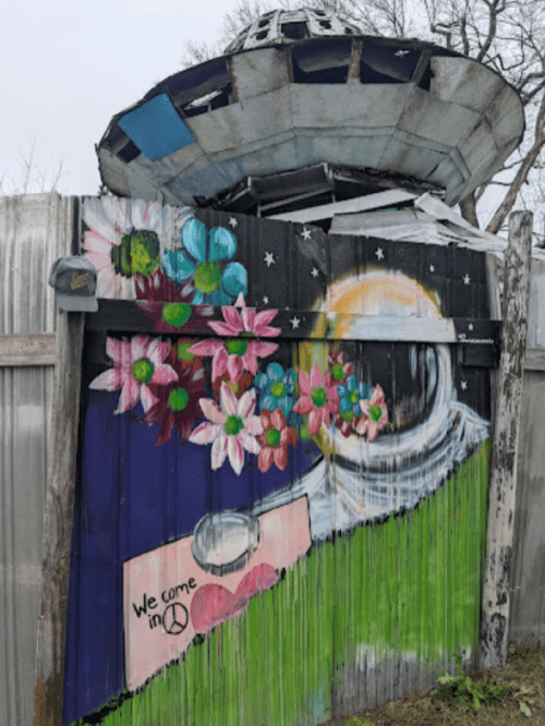 A colorful mural featuring a UFO, flowers, and the phrase "We come in peace" on a wooden fence.