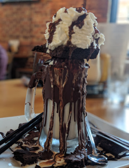 A tall glass filled with milkshake, topped with whipped cream and chocolate sauce, surrounded by brownie pieces.