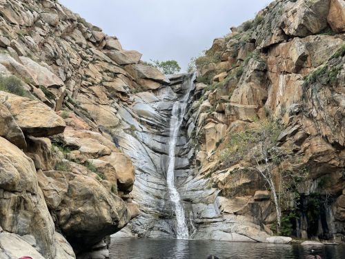 A serene waterfall cascading down rocky cliffs into a calm pool, surrounded by rugged terrain and sparse vegetation.