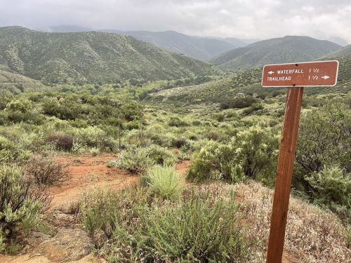 A trail sign points towards a waterfall and trailhead, surrounded by lush green hills and cloudy skies.
