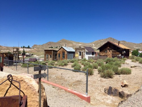 Abandoned wooden buildings in a desert landscape, surrounded by sparse vegetation and mountains under a clear blue sky.
