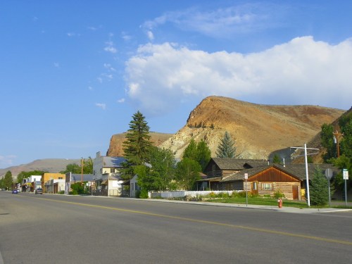 A quiet street lined with rustic buildings and trees, set against a backdrop of rocky hills and a blue sky.