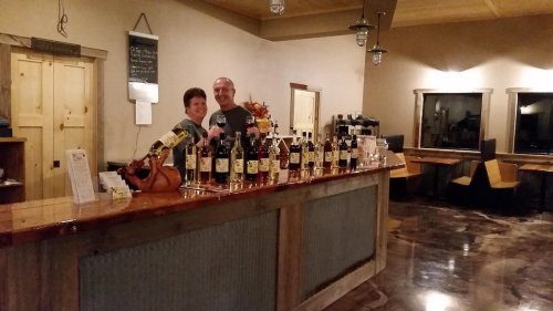 A couple stands behind a bar filled with various bottles, in a cozy, warmly lit tasting room.