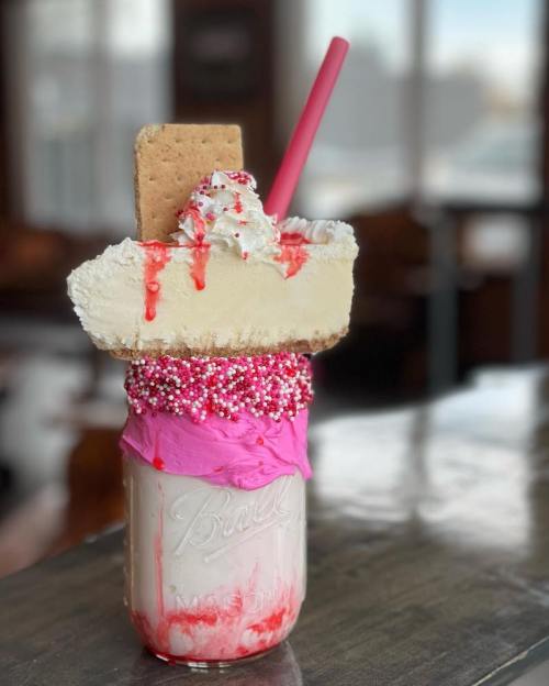 A tall milkshake in a jar topped with whipped cream, a graham cracker, pink frosting, and colorful sprinkles.