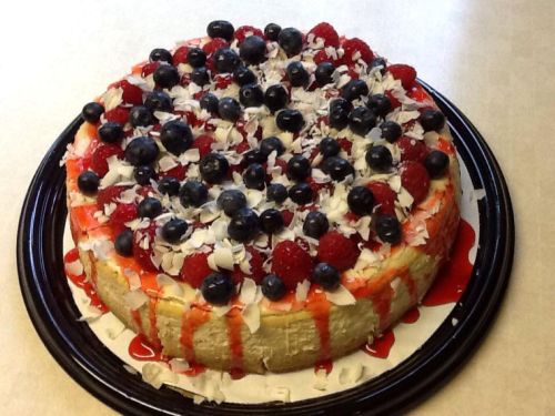 A round cheesecake topped with blueberries, raspberries, shredded coconut, and a drizzle of red sauce.