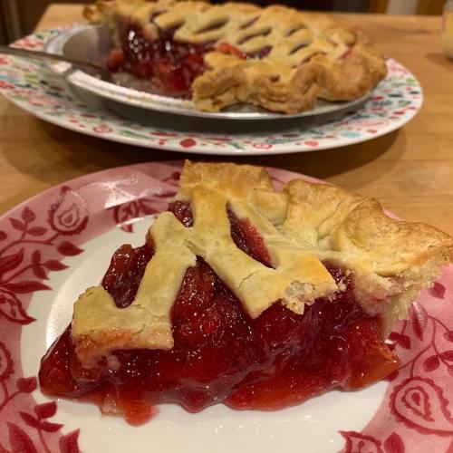 A slice of cherry pie on a decorative plate, with a whole pie in the background on a patterned dish.