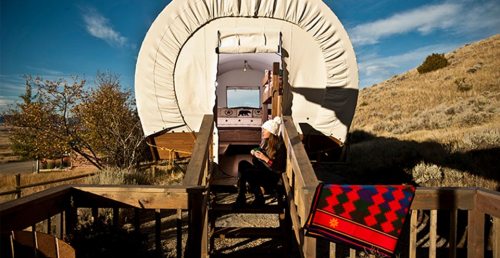 A covered wagon-style accommodation with wooden steps, surrounded by grass and hills, and a colorful blanket draped nearby.