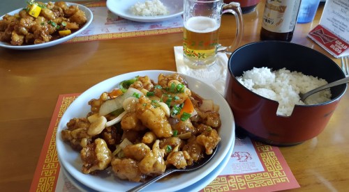 A plate of sweet and sour chicken with vegetables, a bowl of rice, and a glass of beer on a wooden table.