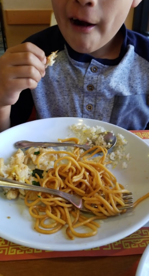 A child is eating from a plate with noodles and rice, holding a piece of food, with utensils on the table.