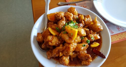 A bowl of sweet and sour chicken with orange slices and green onions, served with a spoon on a wooden table.