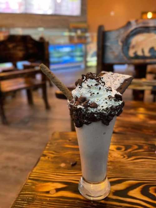 A tall glass of chocolate milkshake topped with whipped cream, chocolate chips, and a cookie, set on a wooden table.