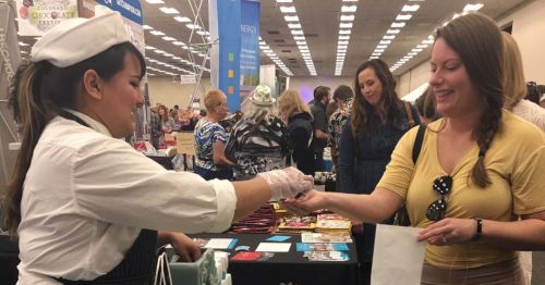 A chef hands a sample to a woman at a busy event, with attendees and booths in the background.