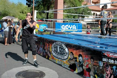 A wrestling ring outdoors with a performer in black attire, while spectators watch nearby under a sunny sky.