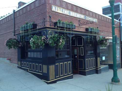 Exterior of Airline Brewing Company, featuring a black and gold facade with hanging flower baskets and a brick wall.