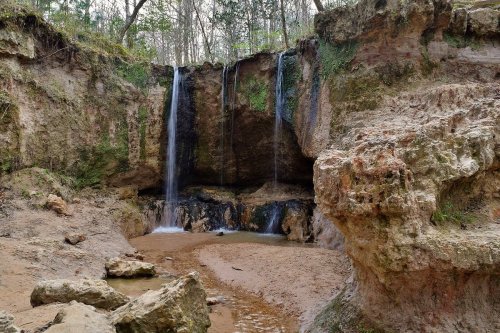 A serene landscape featuring two waterfalls cascading into a rocky, sandy area surrounded by lush greenery.