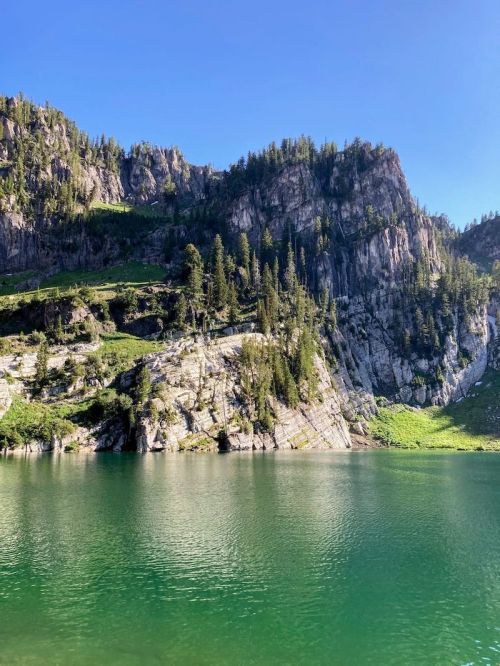 A serene lake surrounded by steep, rocky cliffs and lush greenery under a clear blue sky.