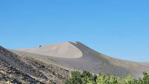 A sandy dune rises against a clear blue sky, surrounded by sparse vegetation and rolling hills.