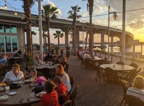 Outdoor dining scene with people at tables, palm trees, and a sunset over a waterfront under a bridge.