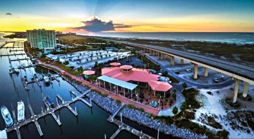 Aerial view of a marina at sunset, featuring boats, a restaurant, and a highway along the coast.