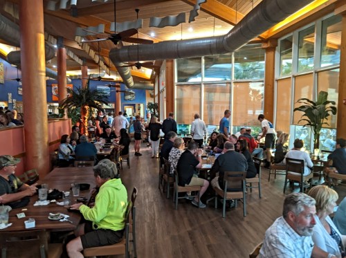 A busy restaurant interior with patrons dining, large windows, wooden beams, and tropical plants.
