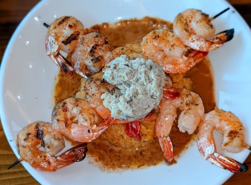 A plate of grilled shrimp skewers on a bed of creamy grits, topped with a dollop of crab meat in a savory sauce.