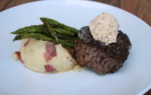 A plate featuring a seasoned beef filet, creamy mashed potatoes with bacon, and grilled asparagus topped with herb butter.