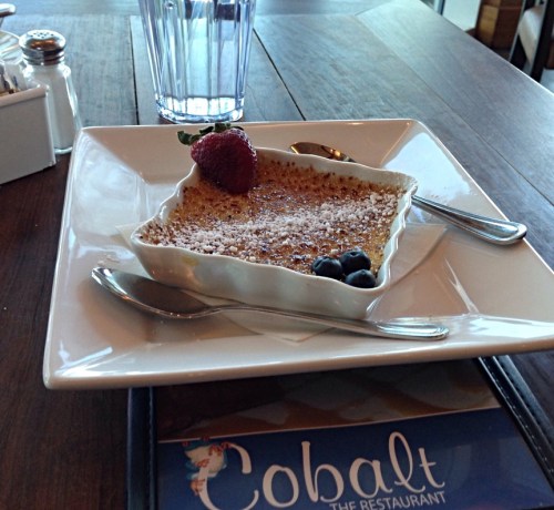 A dessert of crème brûlée in a white dish, garnished with a strawberry and blueberries, on a wooden table.