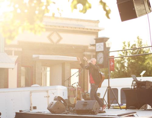 A musician performs on stage with a guitar, surrounded by instruments and a bright, sunny backdrop.