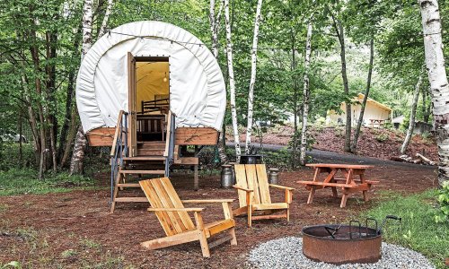 A cozy glamping site with a circular tent, wooden chairs, a picnic table, and a fire pit surrounded by trees.