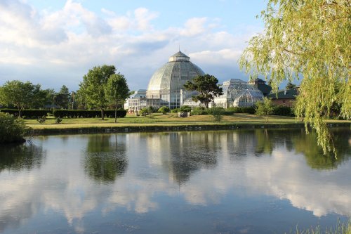 A glass conservatory reflected in a calm lake, surrounded by lush greenery and a cloudy blue sky.
