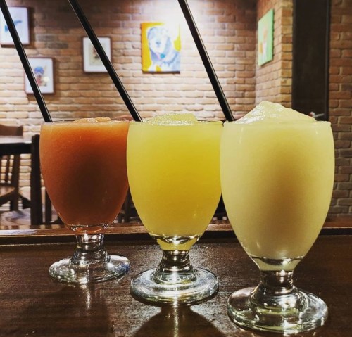 Three colorful frozen drinks in elegant glasses, each with a straw, set against a brick wall and artwork.