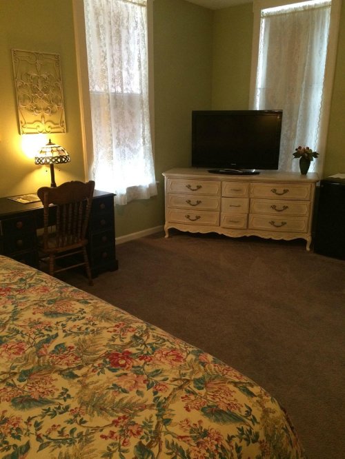 Cozy bedroom with floral bedding, a dresser, TV, desk, and soft lighting from a lamp and windows with lace curtains.