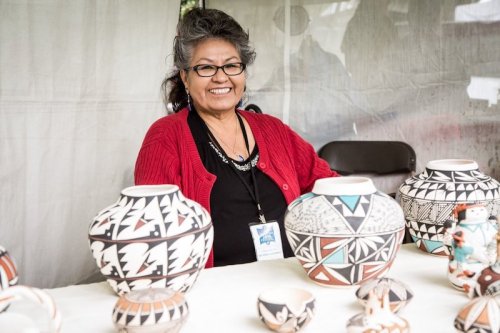 A smiling woman in a red cardigan sits behind a table displaying traditional pottery.