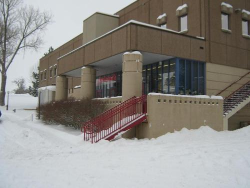 A building entrance with red stairs, surrounded by snow-covered ground and trees, on a winter day.