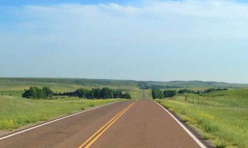 A long, straight road stretches through green fields under a clear blue sky.