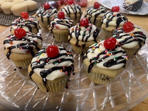 A platter of cupcakes topped with frosting, chocolate drizzle, sprinkles, and cherries.