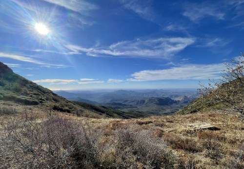 A panoramic view of mountains and valleys under a bright blue sky with scattered clouds and the sun shining above.