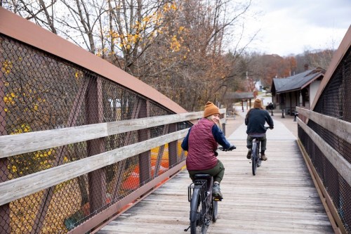 Two people ride bicycles across a wooden bridge surrounded by autumn trees.