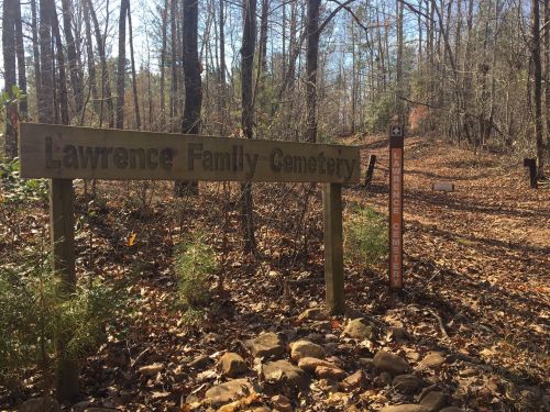 Sign for Lawrence Family Cemetery surrounded by trees and fallen leaves on a wooded path.