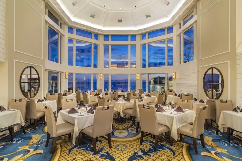 Elegant restaurant interior with large windows, soft lighting, and neatly arranged tables overlooking a scenic view.