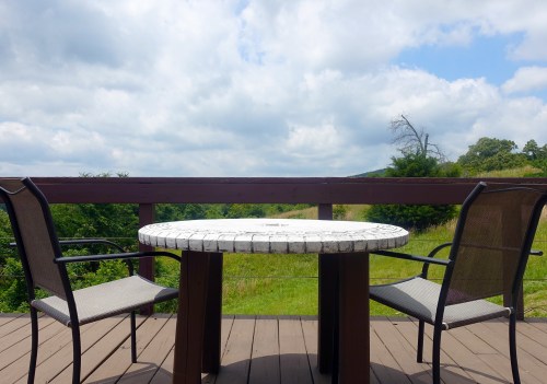 A round table with two chairs on a deck, overlooking a green landscape under a cloudy sky.