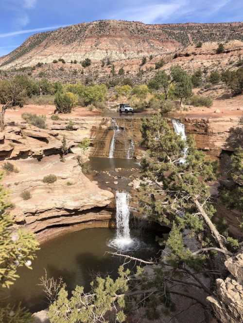 A scenic view of waterfalls cascading into a pool, surrounded by rocky terrain and greenery, with a vehicle in the background.