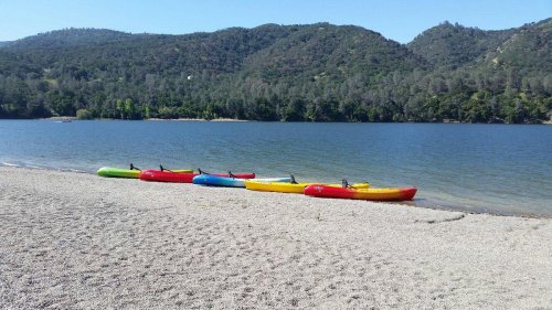 Colorful kayaks lined up on a sandy beach by a calm lake, surrounded by green hills and clear blue skies.