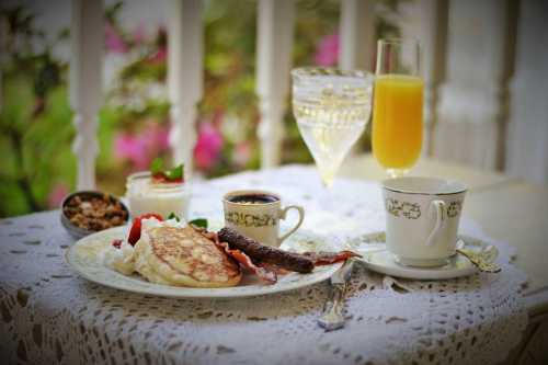 A beautifully arranged breakfast with pancakes, bacon, fruit, coffee, and a glass of orange juice on a lace tablecloth.