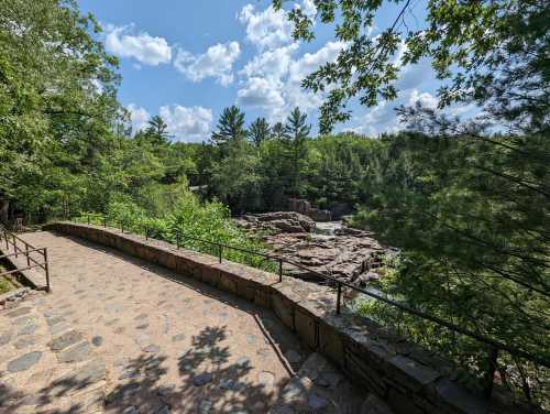 A stone pathway winds through lush greenery, leading to a rocky area under a bright blue sky with fluffy clouds.