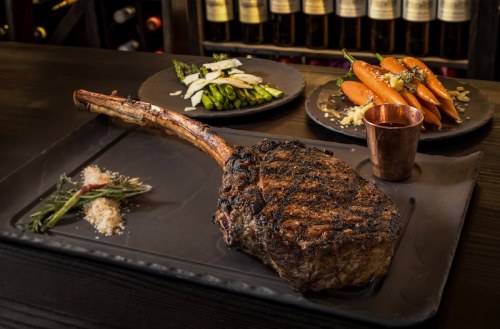 A beautifully cooked tomahawk steak on a black plate, accompanied by asparagus, carrots, and a copper cup.